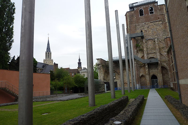 De voormalige St.-Trudo Abdij te Sint-Truiden. De toren van de Abdij staat rechtvoor. De metalen palen symboliseren de oorspronkelijke constructie. Het portiek is aan de linkerzijde van de Abdijtoren zichtbaar. De foto is genomen vanuit het pad dat afdaalt naar de crypte. Aan de horizon zijn de torens van het stadhuis (midden) en de O.L.Vrouwekerk (links) zichtbaar.